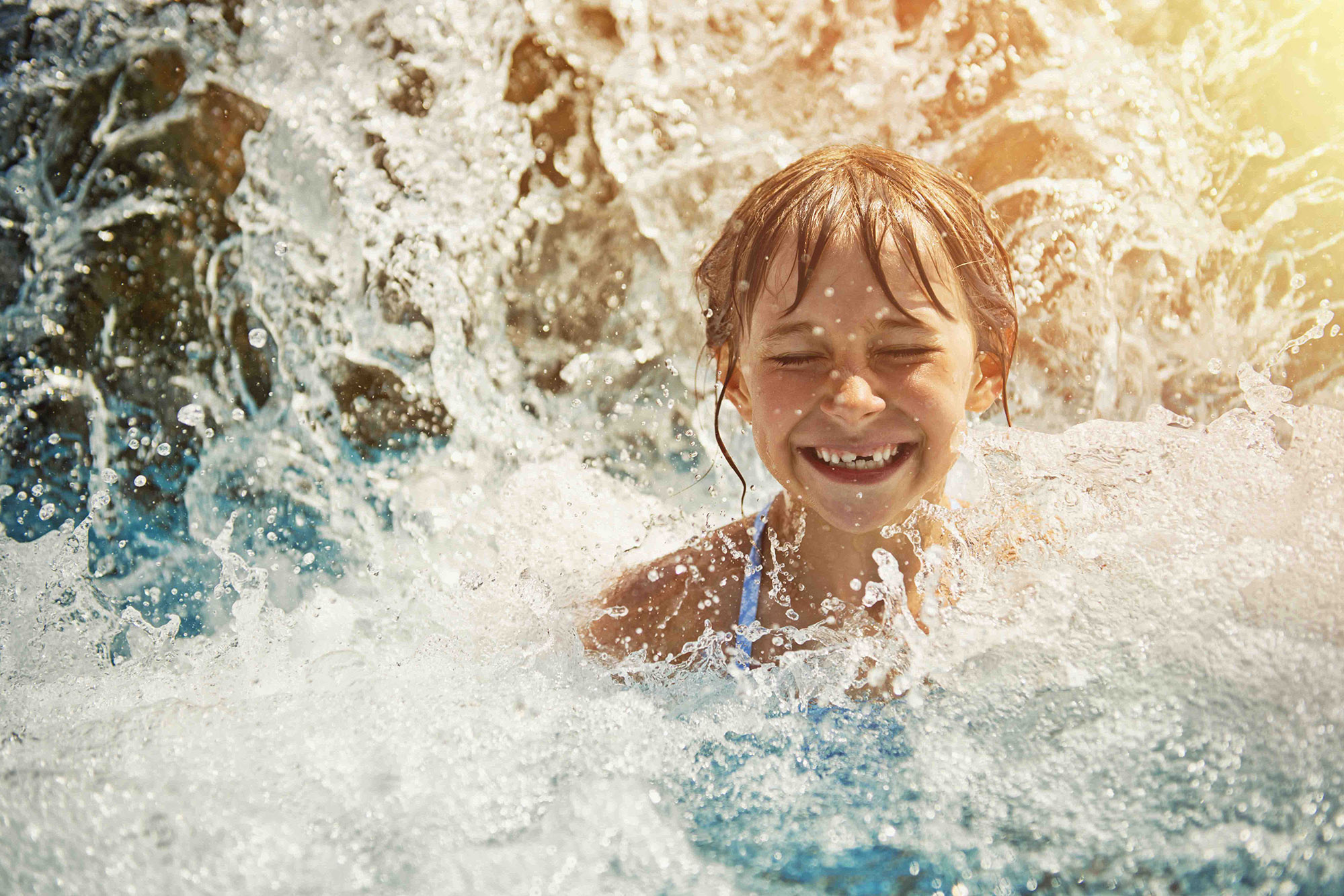 Little girl in waterpark pool being splashed by waterfall. Sunny summer day.