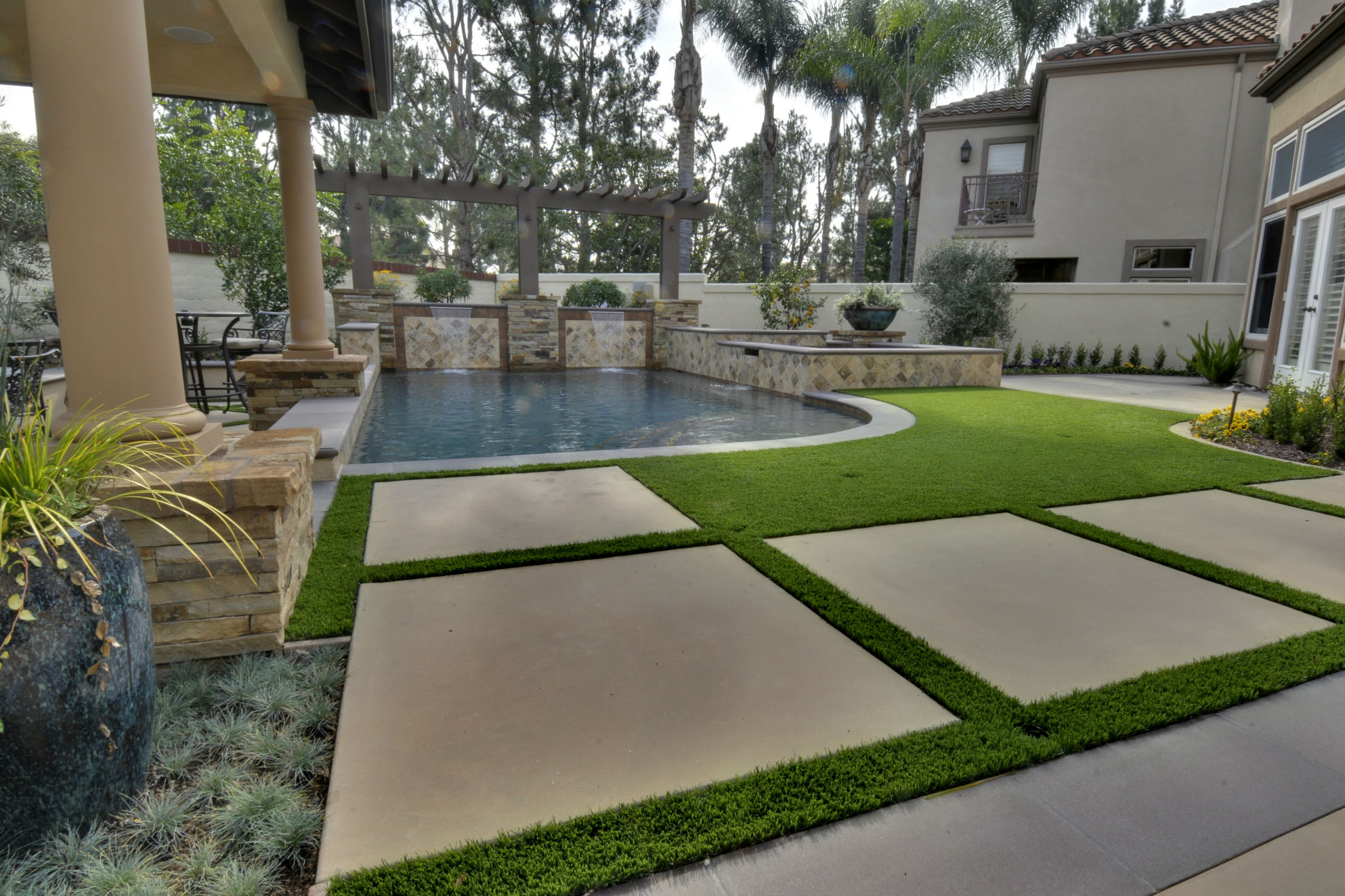 Synthetic grass surrounds the stepping pads and pool area of this backyard project by Conscious Environments, an important feature to the homeowner, who was interested in a low-maintenance, drought-friendly alternative. Ceramic pottery and Mediterranean plantings provide balance to the worry-free greenery.