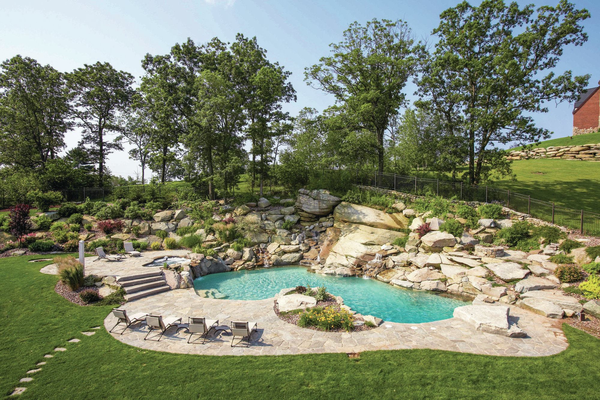 From the house, the homeowners can see a series of natural looking “waterfalls” that feed into the pool. The deep end of the pool, at right, features a diving rock rather than a board. The flat rock “steps” to the right of the diving rock lead up to a fire pit. “It’s a family with five boys,” says Giannamore, “and we knew they’d be scrambling all over the rocks anyway, so we wanted to at least allow easy access to the fire area, which we knew they’d be using a lot — hopefully without twisting an ankle to get there.”