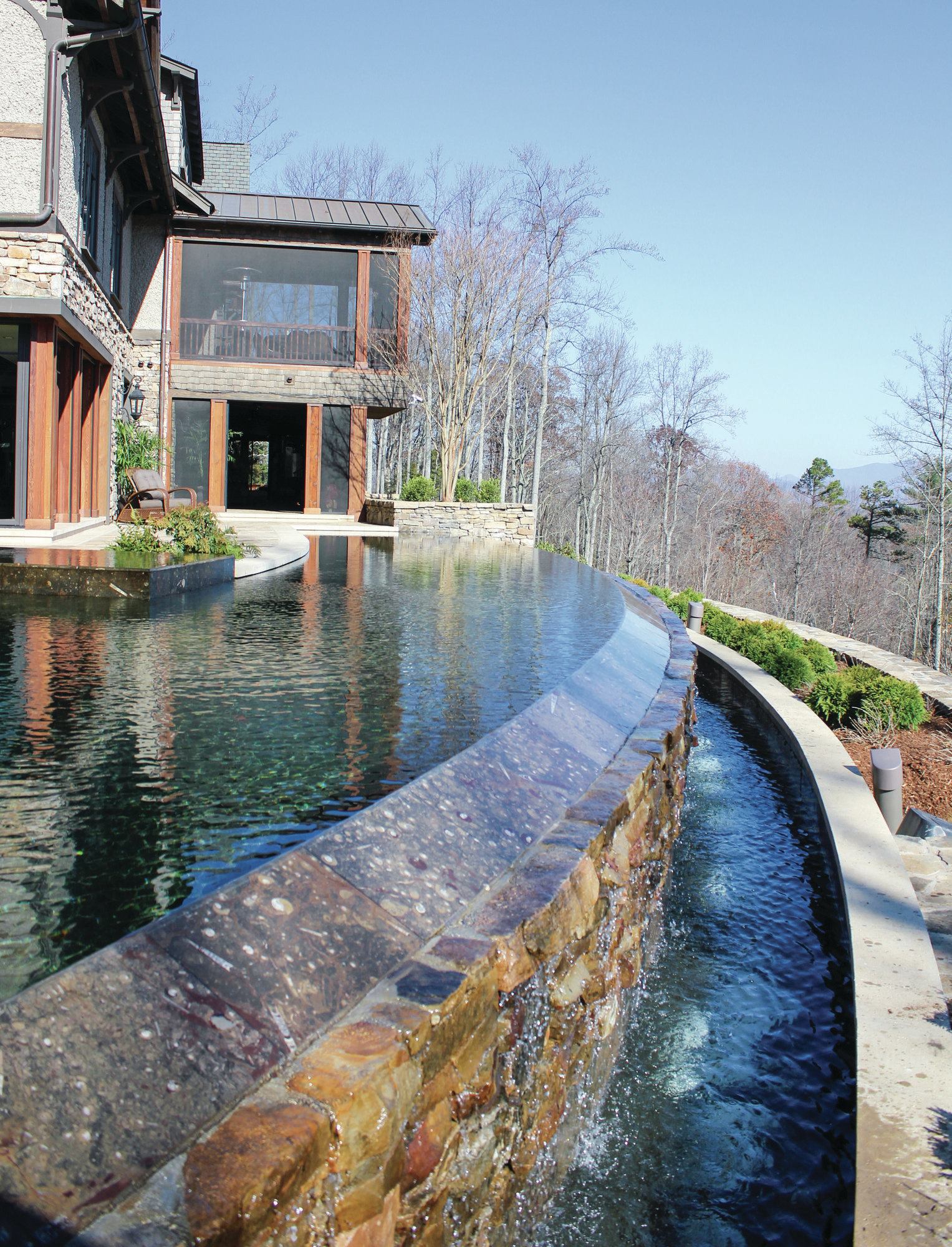 Sweeping View: The 77-foot-long vanishing-edge pool forms a sweeping arc connecting the main house with the guest house, seen in the top image. A black pebble finish adds to the reflective quality and adds a modern touch, while the Tennessee fieldstone deck ties with the home’s rustic materials.