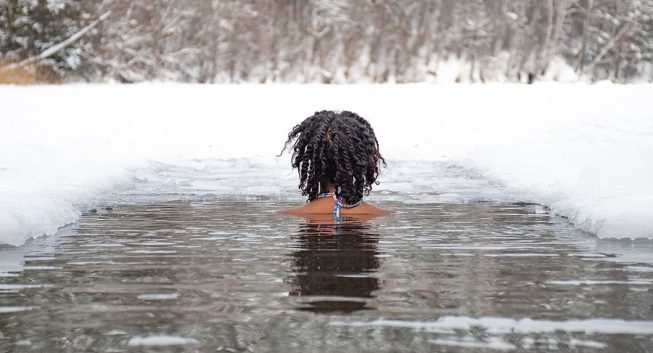 Person with curly hair standing in partially frozen water, surrounded by snow and ice.