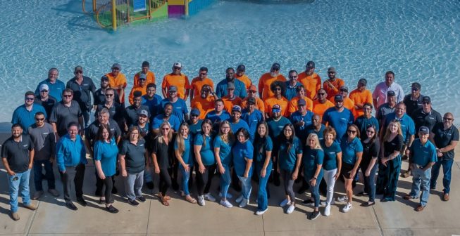 A group of diverse adults in blue and orange shirts stand smiling by a water park pool with slides in the background, conveying a cheerful atmosphere.