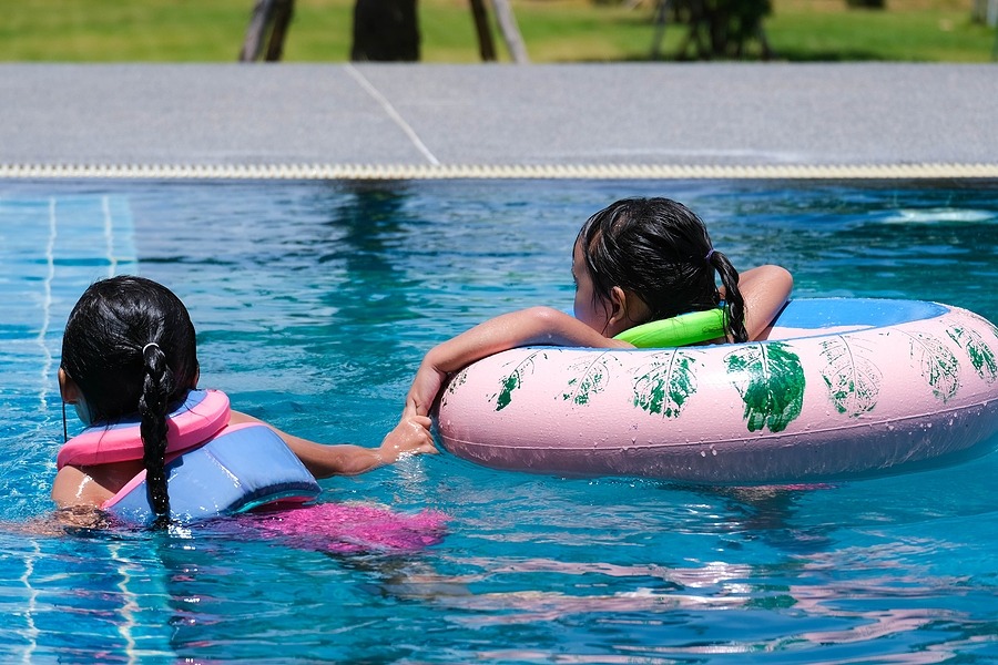 Two children swim in a bright blue pool; one wears a pink life jacket while swimming, and the other floats on a large pink inflatable ring.