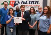 Seven adults pose with a safety achievement certificate in front of a North Carolina Department of Labor NCDOL banner.