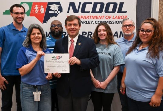 Seven adults pose with a safety achievement certificate in front of a North Carolina Department of Labor NCDOL banner.