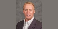 Head-and-shoulders portrait of a man with short reddish-blond hair wearing a dark blazer and light button-down shirt against a gray background.