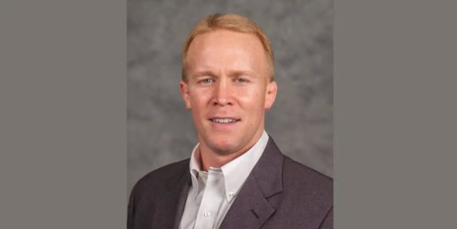 Head-and-shoulders portrait of a man with short reddish-blond hair wearing a dark blazer and light button-down shirt against a gray background.