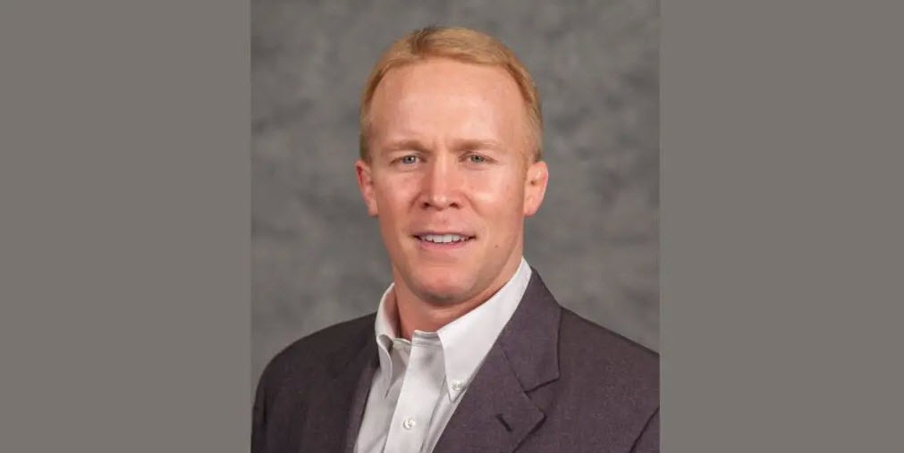 Head-and-shoulders portrait of a man with short reddish-blond hair wearing a dark blazer and light button-down shirt against a gray background.